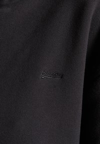 Musta collegekangas, jossa on sileä pinta, ja jossa on kohopainettu "SuperDry" logo hieman tummemmassa sävyssä.