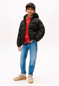 Črna puhovka s kapuco, rdeča pletena majica, svetlo modri kavbojki in bež superge; opazna prešita tekstura in sproščen kroj.