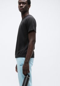 Μαύρο κοντομάνικο μπλουζάκι, ανοιχτό μπλε φόρμα με λογότυπο Puma. Το μοντέλο κρατάει μια μαύρη θερμός νερού, στέκεται μπροστά σε λευκό φόντο.