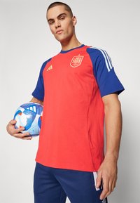 Punane ja sinine lühikeste varrukatega spordipliid, millel on Hispaania embleem ja kolm valget triipu õlgadel. Hoia sinist mustriga jalgpalli.