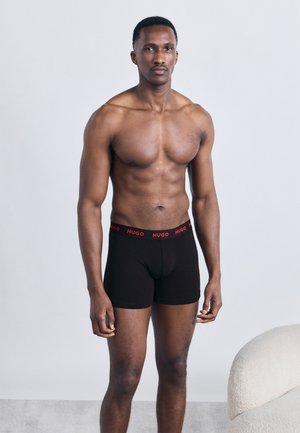 Muskulózny muž stojaci v čiernych boxerkách Hugo na jednoduchom sivom pozadí pri bielom textúrovanom kresle.