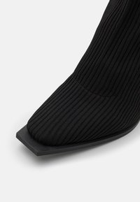 Zwarte gebreide enkelboot met een spitse neus, geribbelde textuur en een stevige vierkante hak. De zool is gemaakt van glad zwart materiaal.