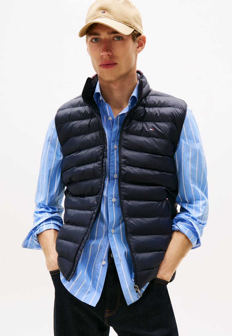 Jovem a usar boné bege, camisa de botões azul às riscas, colete acolchoado preto e jeans escuros, em pé com as mãos nos bolsos.