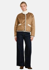 Giacca in suede marrone con rifiniture in shearling bianco, caratterizzata da un colletto rotondo, chiusura con bottoni e due tasche frontali; abbinata a jeans neri a gamba larga.