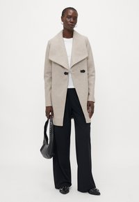 Beige wollen blend jas met een sjaalkraag, twee zwarte knopen en een gestroomlijnde silhouette. Model combineert het met zwarte broeken en een handtas.