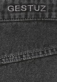 Tissu en denim noir avec texte brodé blanc "GESTUZ" au-dessus d'une couture, montrant la texture détaillée et les motifs de fil.