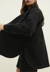 Chaqueta de denim negra oversized con dobladillo curvado, hombros caídos y un ajuste relajado. Presenta costuras visibles y un diseño minimalista.