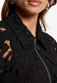 Veste à col noir avec de la dentelle florale texturée, fermeture éclair à l'avant et détails découpés, mettant en valeur des motifs intriqués et un matériau léger.