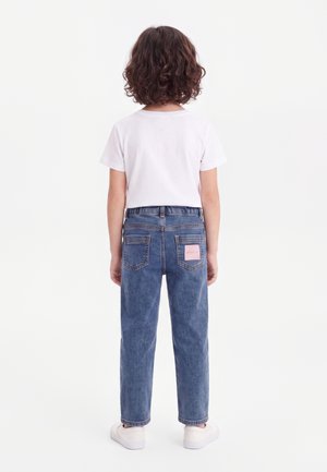 Kind mit lockigem dunklem Haar steht mit dem Rücken zur Kamera, trägt ein schlichtes weißes T-Shirt, blaue Jeans mit Gesäßtaschen und weiße Turnschuhe.