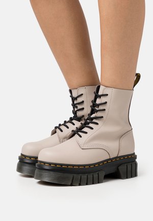 Beige leather lace-up boots with black thick ridged soles and yellow stitching worn on bare legs, shown from side on white background.