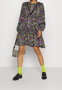 Černé květinové šaty s dlouhými rukávy. Jasné multicolor vzory, krajkové detaily na výstřihu. Kombinované s neonově zelenými ponožkami a pruhovanou taškou.