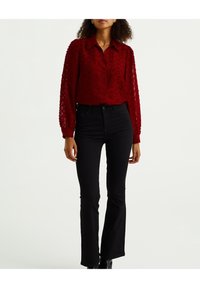 Rote, strukturierte Bluse mit Kragen und durchgehender Knopfleiste; kombiniert mit schwarzen, weit geschnittenen Jeans. Der Stoff weist ein florales Muster und durchsichtige Ärmel auf.