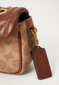 Brown textured leather handbag featuring a quilted top, gold hardware, and a brown leather tag attached by a beaded chain.