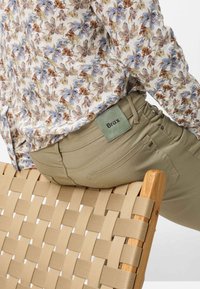 Personne portant une chemise fleurie et un pantalon beige avec l'étiquette "Brax", assise sur une chaise tressée beige avec un cadre en bois.
