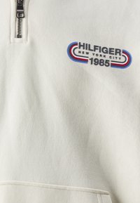 Felpa zip in crema chiaro con texture a coste. Presenta un logo ricamato "HILFIGER NEW YORK CITY 1985" in blu, rosso e grigio.