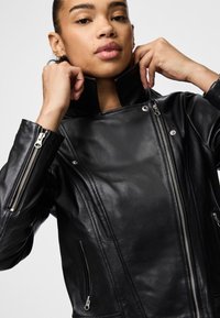 Must mustrõngaga must nahast mootorrattajope, mis sisaldab hõbedast riistvara ja diagonaalseid tõmblukuga taskuid. Sile tekstuur ja sobituv disain.