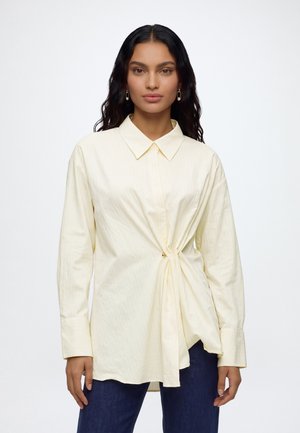 Femme aux longs cheveux foncés portant un chemisier à manches longues couleur crème avec un nœud latéral et un pantalon bleu foncé, debout sur fond blanc.