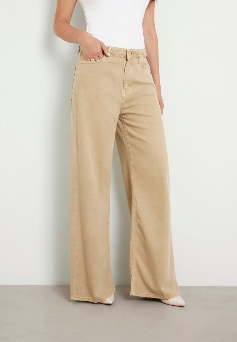 Pantalones de pierna ancha beige con cintura alta, hechos de una suave tela de algodón. Cuenta con bolsillos delanteros y un cierre clásico con botón. Textura suave.