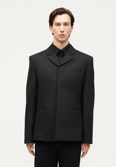 Jeune homme aux cheveux foncés portant une veste de costume noire surdimensionnée et une chemise noire, debout devant un fond clair uni.