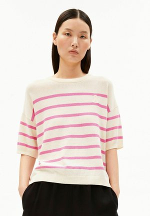 Jonge vrouw met stijl donker haar, gekleed in een wit kort mouwtruitje met horizontale roze strepen en zwarte broek, handen in de zakken.