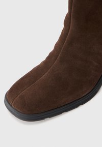 Botte en suède marron, haute jusqu'au genou, avec une texture lisse, un bout rond et une semelle en caoutchouc noire. Détails de couture sur le devant.