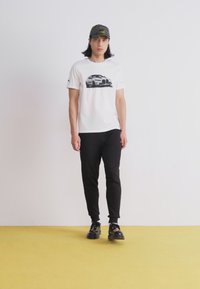 Hvid t-shirt med bilgrafik, parret med sorte slim-fit bukser og sorte sko. Model iført en camouflagekasket, stående på gulv i gul farve.