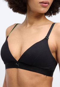 Žena oblečená v čiernej športovej podprsenke Puma s nastaviteľnými ramienkami a jemným logom, zobrazená od hrudníka až po spodnú časť tváre na jednoduchom pozadí.