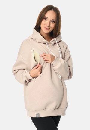 Frau trägt einen beigen Hoodie mit Still-Reißverschluss und öffnet die Seitennaht, um den hellen Still-BH darunter zu zeigen.