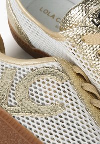 Mřížkové tenisky s bílým základem a zlatými akcenty, texturovaným zlatým potahem, béžovými tkaničkami a hnědou gumovou podrážkou. Viditelný vzorovaný design.