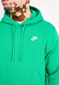 Sudadera verde de Nike con un bolsillo canguro frontal, capucha con cordones y logo blanco en el pecho. Tejido suave con una textura lisa.
