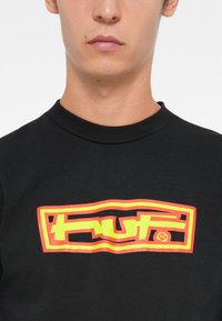 Black cotton t-shirt featuring a bold, multicolored logo in yellow and orange, with a smooth texture and short sleeves.