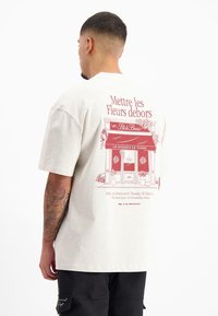 Beige katoenen t-shirt met korte mouwen en een achterafbeelding met een rode bloemenwinkelillustratie en tekst, inclusief "Mettre les Fleurs debors."