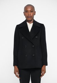 Μαύρο διπλόπετο παλτό από μάλλινο ύφασμα, με μεγάλα πέτα, μαύρα κουμπιά και δύο πλαϊνές τσέπες, που φοριέται πάνω από ένα λευκό πουκάμισο με γιακά.