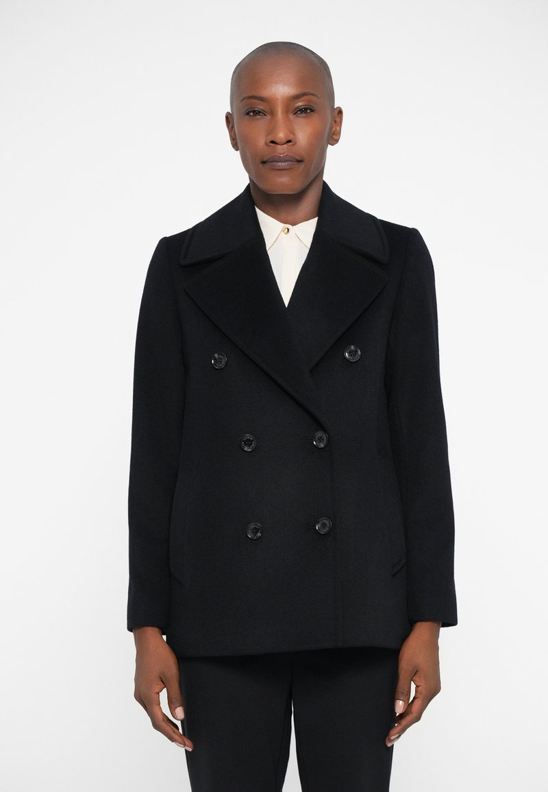 Μαύρο διπλόπετο παλτό από μάλλινο ύφασμα, με μεγάλα πέτα, μαύρα κουμπιά και δύο πλαϊνές τσέπες, που φοριέται πάνω από ένα λευκό πουκάμισο με γιακά.