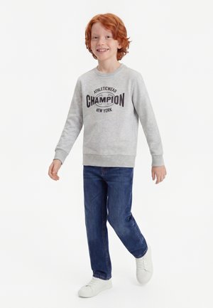 Rødhåret dreng smilende, iført en grå sweatshirt med teksten "Champion", blå jeans og hvide sneakers, går mod en hvid baggrund.