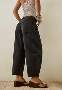 Vrouw in losse zwarte cropped jeans, een doorschijnend patroon top, chunky armbanden en bruine wedge sandalen, staande op een betegelde vloer.