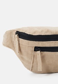 Marsupio in corduroy beige con due cerniere nere e tessuto texture. Presenta una forma curvata e bordi arrotondati per un design compatto.