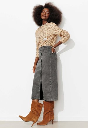 Femme aux cheveux afro volumineux portant un chemisier à fleurs, une jupe midi en jean gris avec une fente à l'avant, des bottes à talons hauts en daim marron et des bijoux en perles.