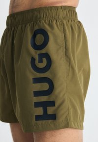Olivovo zelené plavky s elastickým pásom a veľkým tmavomodrým logom "HUGO" na ľavej strane. Hladký materiál so bočnými vreckami.