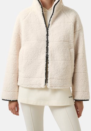 Persona que lleva una chaqueta de forro polar sherpa color crema sobre una sudadera con capucha y leggings a juego en color crema, de pie frente a un fondo sencillo.