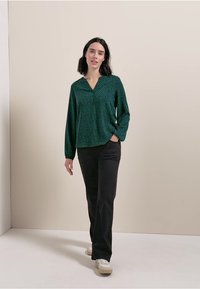 Grün gemusterte Bluse mit V-Ausschnitt, langen Ärmeln und lockerer Passform, kombiniert mit schwarzen Hosen und weißen Sneakern. Glatte Stoffstruktur.