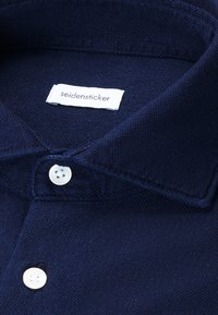 Camicia in cotone blu navy con colletto strutturato, dotata di etichetta bianca e bottoni bianchi. Texture liscia e elementi di design classici.