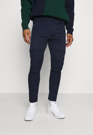 Homme portant un pantalon cargo bleu marine, des baskets blanches et un pull à blocs de couleur verts et bleu marine, debout devant un fond uni.