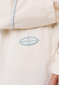 Beige lichtgewicht jas met een zachte textuur, blauwe stiksels en een ronde blauwe logo met de letters "PXP" duidelijk weergegeven.