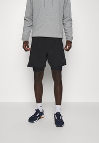 Černé sportovní šortky s hladkým povrchem, nošené přes těsné šortky. V kombinaci se šedou mikinou a námořnickými sportovními botami.