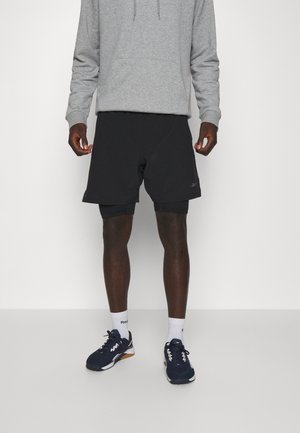 Pantaloni corti sportivi neri con una texture liscia, indossati sopra pantaloni attillati. Abbinati a un felpa grigia e scarpe sportive blu navy.