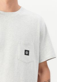 Gray cotton t-shirt featuring a crew neck, short sleeves, and a chest pocket with a black logo patch. Soft texture with a simple design.