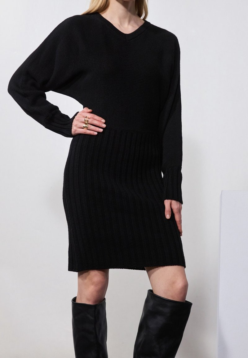 Femme portant une robe noire en maille côtelée à manches longues avec un col en V et des bottes en cuir noir jusqu’aux genoux, accessoirisée de bagues en or.