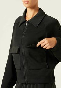 Schwarze Zip-Jacke aus glattem Stoff. Verfügt über einen breiten Kragen, lange Ärmel und zwei Fronttaschen. Einfaches, modernes Design.