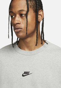 Jauns cilvēks ar pīts matiem, ģērbts gaiši pelēkā džemperī ar mazu melnu Nike logotipu uz krūtīm, skatās uz sānu.